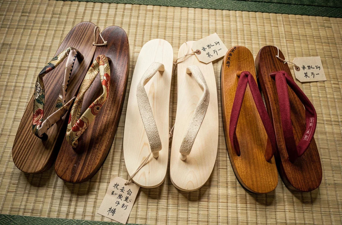 Assortiment de trois paires de geta artisanales japonaises en différents bois et matières de sangles, disposées sur un tatami avec des étiquettes calligraphiées